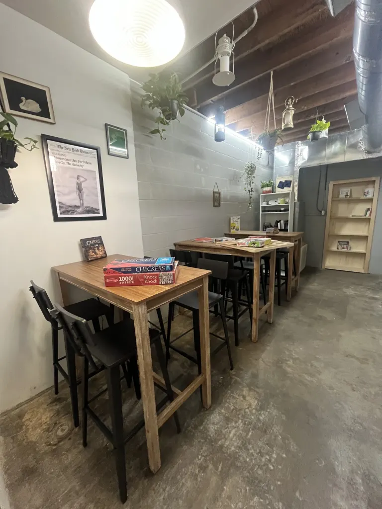 Narrow room with wooden high-top tables, black bar stools, framed prints, a bookshelf and a paper lantern pendant under a wood-beam ceiling