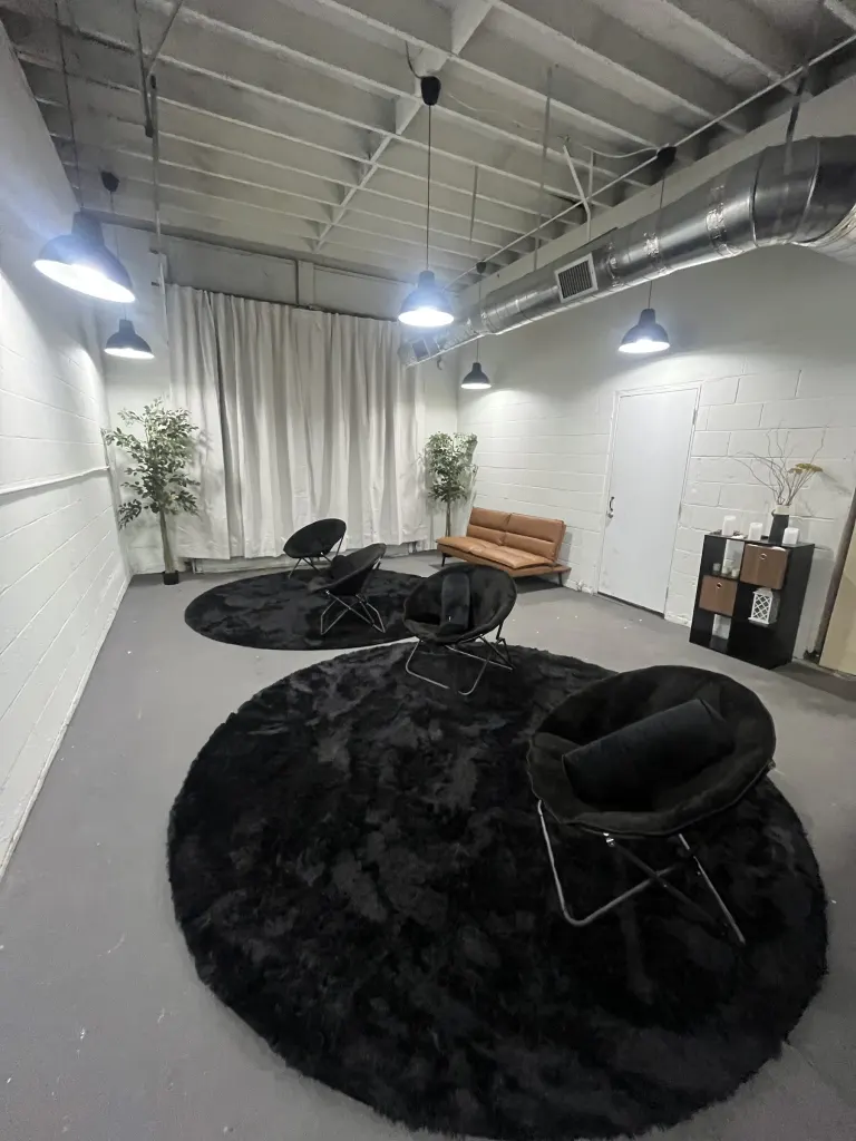 Industrial studio room with white brick walls, black pendant lights, sheer white curtains, round black shag rugs and black lounge chairs