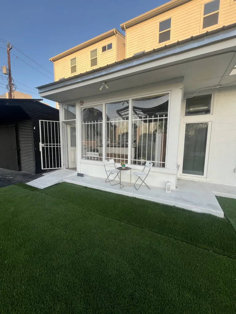Exterior of HTX Bodega at golden hour: a green turf yard leading to a patio with a white bistro table and chairs beside large windows